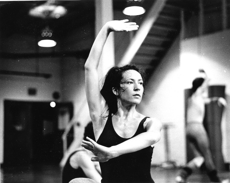 a black and white photo of a young white woman with dark hair in a sleeveless black leotard dancing in a studio looking pensive, the background the out of focus shows 2 other figures dancing behind her